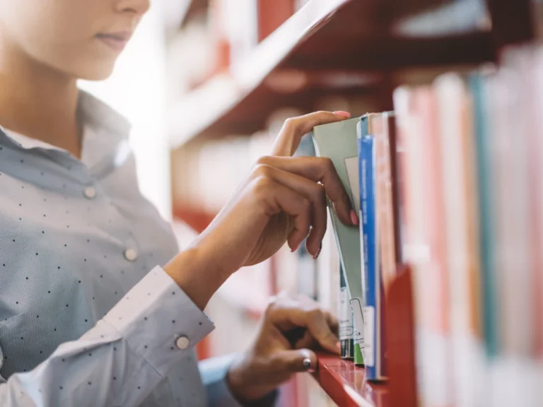 A woman browsing a library bookshelf, pulling a book from the shelf.