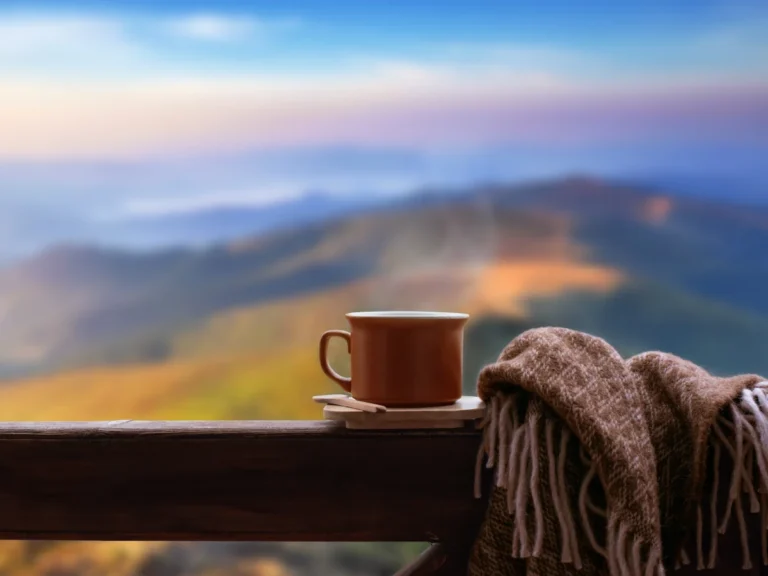 A steaming mug of coffee and a wool blanket on a wooden railing overlooking a mountain landscape at sunrise.