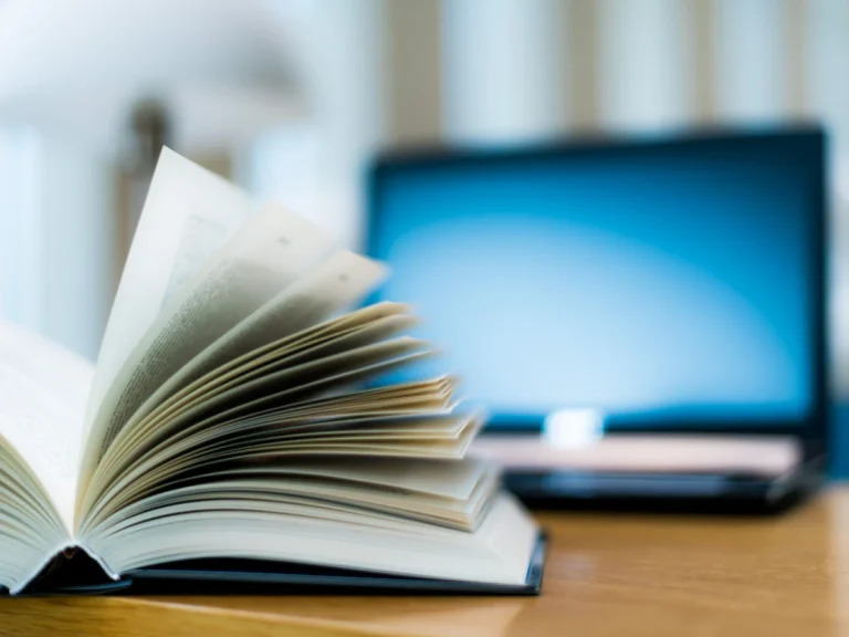 An open book with fanned pages on a wooden desk, with a laptop blurred in the background.