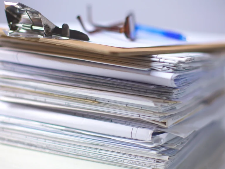 A tall stack of paper documents with glasses and a pen resting on top.