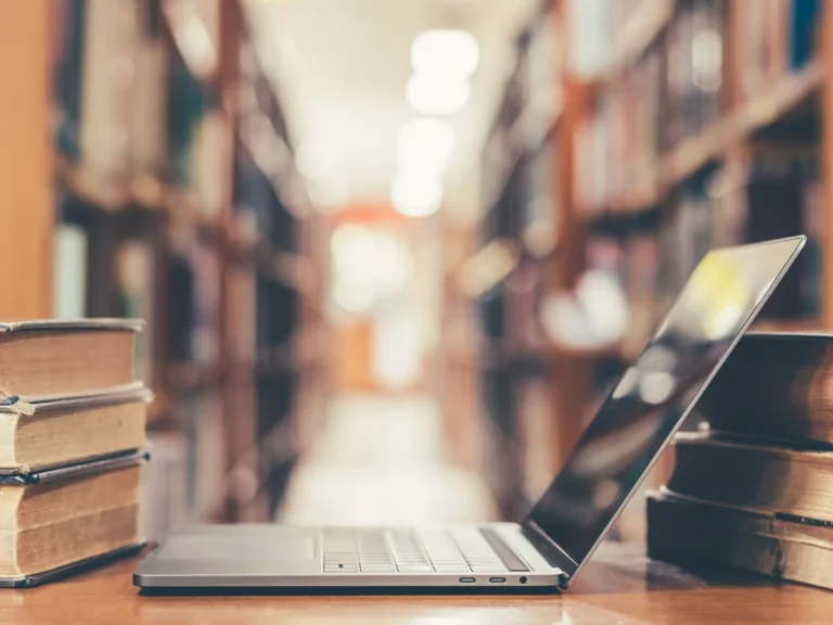 An open laptop on a wooden table between stacks of books, with blurred library shelves in the background.
