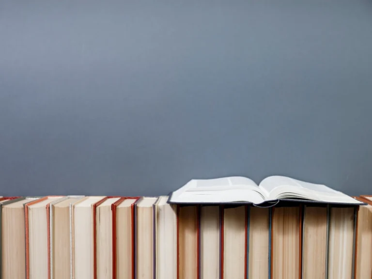 An open book resting on top of a row of upright books against a gray background.